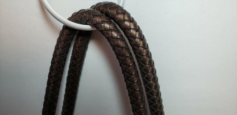May include: Close-up of a brown braided leather strap, looped over a white circular object. The leather has a metallic sheen. The background is a plain white surface. The strap appears to be part of a bag or accessory.