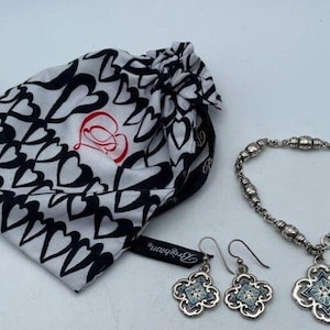 May include: A silver bracelet with a charm in the shape of a four-leaf clover. The charm has a blue and white enamel design. The bracelet is packaged in a white and black bag with hearts printed on it. The bag has the text "Betsey Johnson" printed on it.