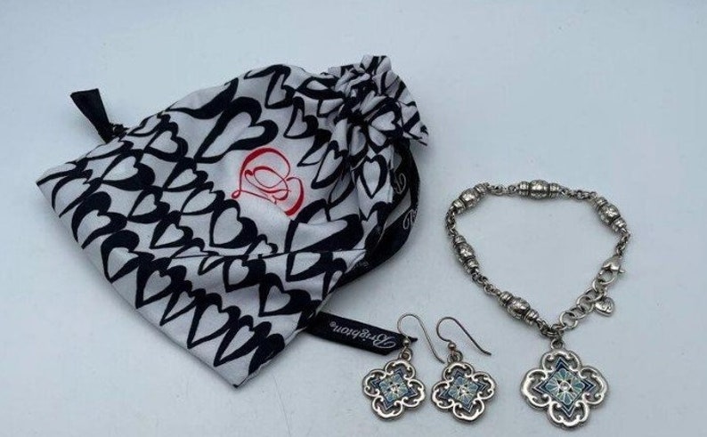 May include: A silver bracelet with a charm in the shape of a four-leaf clover. The charm has a blue and white enamel design. The bracelet is packaged in a white and black bag with hearts printed on it. The bag has the text "Betsey Johnson" printed on it.