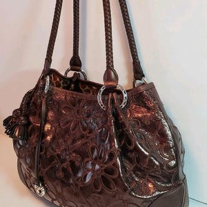 May include: A brown leather handbag with a floral embossed design. The bag features braided handles, silver-tone hardware, and a tassel detail. The interior lining has a leopard print.