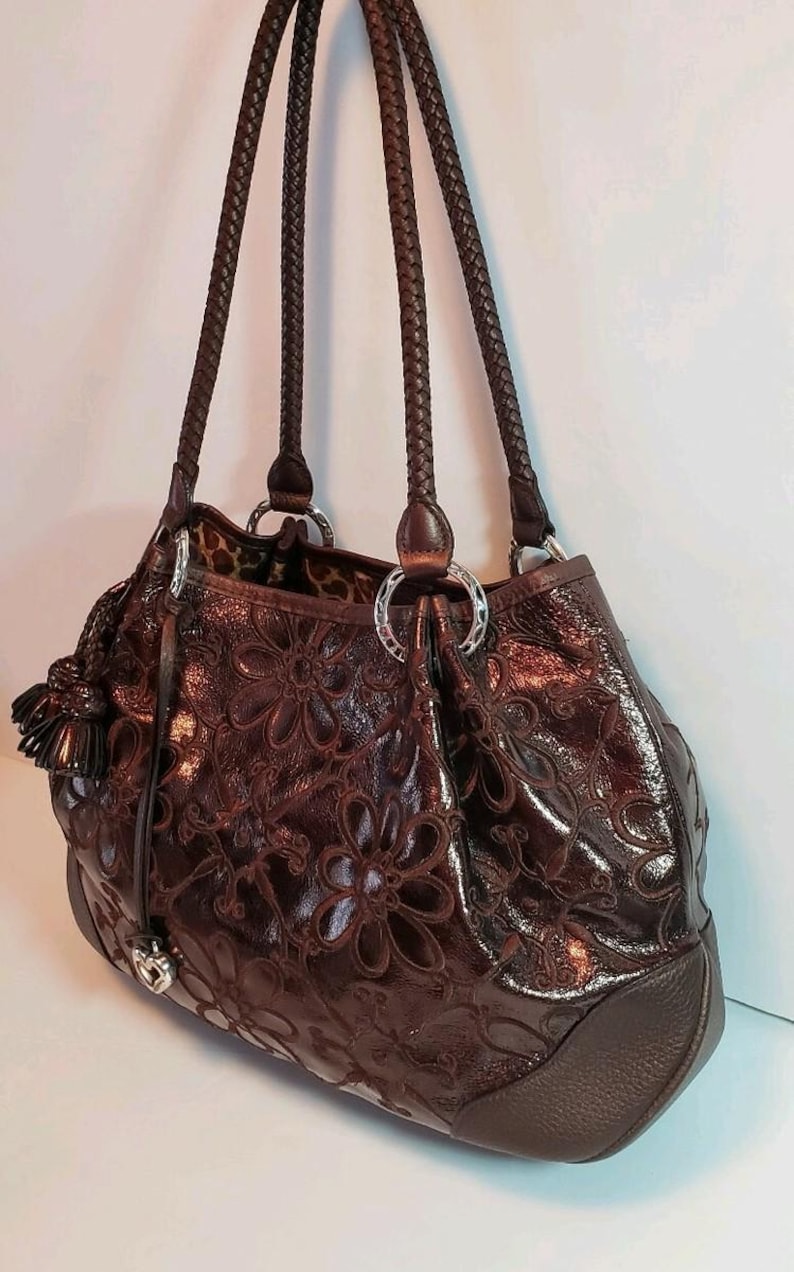 May include: A brown leather handbag with a floral embossed design. The bag features braided handles, silver-tone hardware, and a tassel detail. The interior lining has a leopard print.