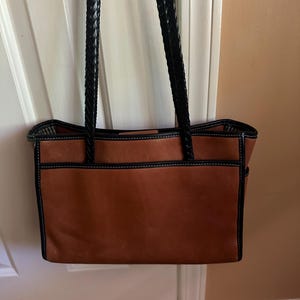 May include: A brown leather tote bag with black braided handles. The bag has black trim and white stitching. The bag is hanging on a white door.