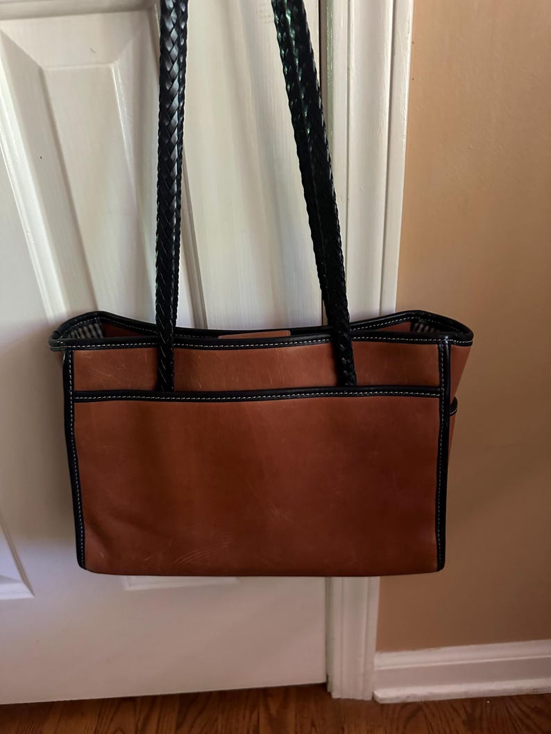 May include: A brown leather tote bag with black braided handles. The bag has black trim and white stitching. The bag is hanging on a white door.