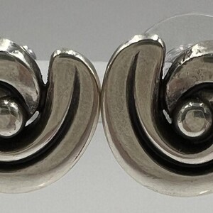 以下が含まれることがあります： シルバーカラーのスタッドピアス2個セット。各イヤリングは、中央に小さな丸いアクセントが付いた渦巻くスパイラルデザインが特徴です。イヤリングは研磨仕上げで、ポストバックが付いています。