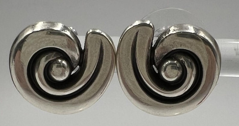 以下が含まれることがあります： シルバーカラーのスタッドピアス2個セット。各イヤリングは、中央に小さな丸いアクセントが付いた渦巻くスパイラルデザインが特徴です。イヤリングは研磨仕上げで、ポストバックが付いています。