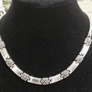 Puede incluir: Collar plateado con cadena de serpiente y eslabones rectangulares. Los eslabones est&aacute;n conectados por elementos decorativos cuadrados. El collar se exhibe en un maniqu&iacute; negro.