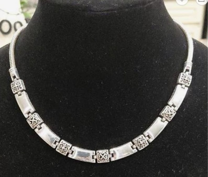 Puede incluir: Collar plateado con cadena de serpiente y eslabones rectangulares. Los eslabones est&aacute;n conectados por elementos decorativos cuadrados. El collar se exhibe en un maniqu&iacute; negro.