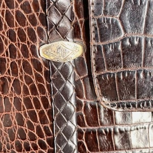 May include: Close-up of a brown leather bag with a crocodile skin texture. The bag features a braided leather strap and a silver-colored clasp with an ornate design. The overall aesthetic is vintage and luxurious.