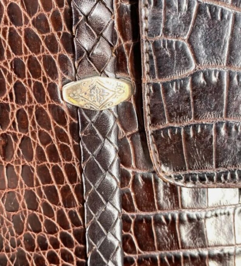 May include: Close-up of a brown leather bag with a crocodile skin texture. The bag features a braided leather strap and a silver-colored clasp with an ornate design. The overall aesthetic is vintage and luxurious.
