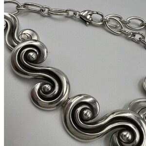 以下が含まれることがあります： 銀色のネックレス。一連の様式化された渦巻きのデザインが特徴です。ハート型の留め金が付いたチェーンが付いています。大胆でエレガントなジュエリーです。