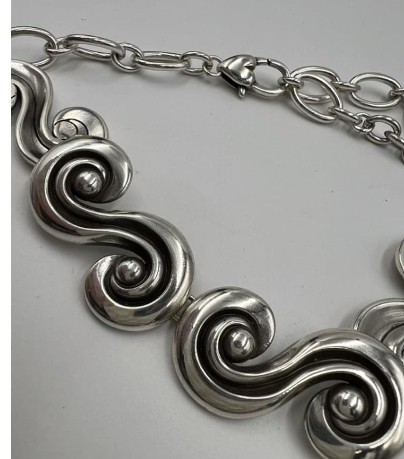 以下が含まれることがあります： 銀色のネックレス。一連の様式化された渦巻きのデザインが特徴です。ハート型の留め金が付いたチェーンが付いています。大胆でエレガントなジュエリーです。