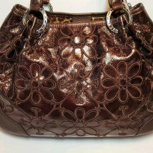 May include: A brown leather handbag with a floral embossed design. The bag features a glossy finish, silver metal rings, and a patterned interior. The floral pattern is repeated across the front and sides of the bag.