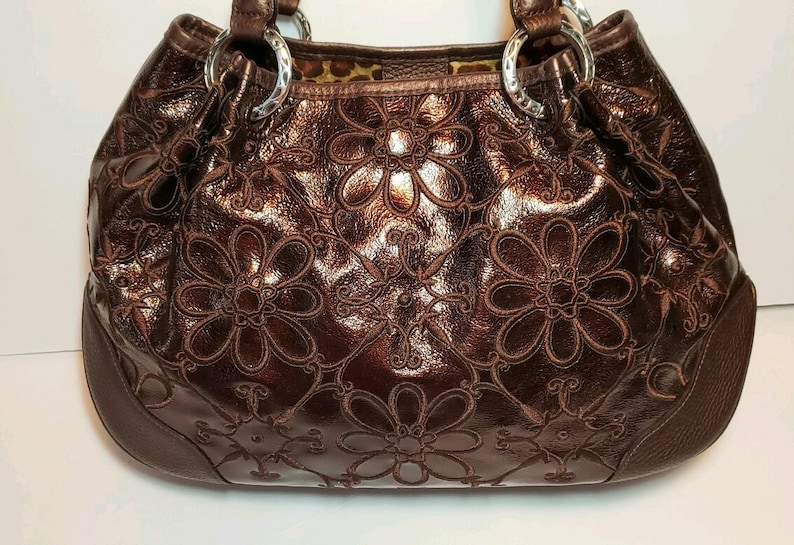 May include: A brown leather handbag with a floral embossed design. The bag features a glossy finish, silver metal rings, and a patterned interior. The floral pattern is repeated across the front and sides of the bag.