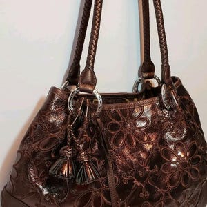 May include: A brown leather handbag with a floral pattern embossed on the exterior. The bag has braided handles, silver-tone rings, and decorative tassels. The bottom of the bag is a darker brown.
