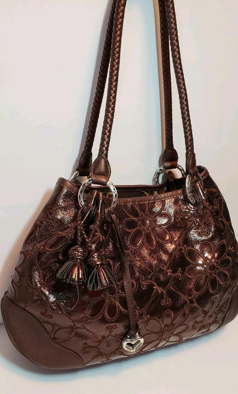 May include: A brown leather handbag with a floral pattern embossed on the exterior. The bag has braided handles, silver-tone rings, and decorative tassels. The bottom of the bag is a darker brown.