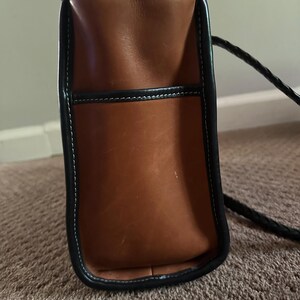 May include: A brown leather handbag with black trim and a braided black strap. The bag has white stitching and a rectangular shape. The leather appears to be worn, giving it a vintage look.