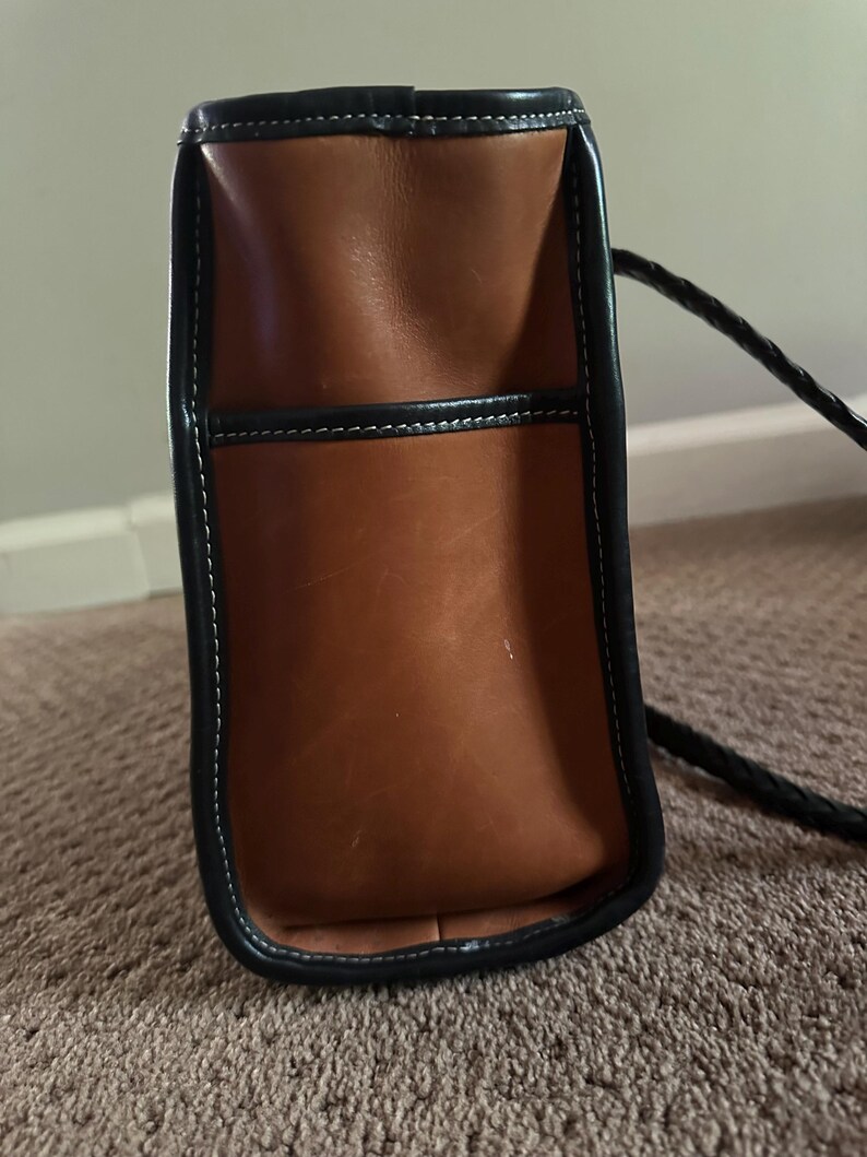 May include: A brown leather handbag with black trim and a braided black strap. The bag has white stitching and a rectangular shape. The leather appears to be worn, giving it a vintage look.
