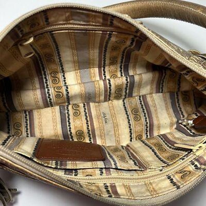 May include: Tan handbag with a patterned interior. The inside features a striped design with decorative swirls and a brown leather tag. The bag has a zipper closure and a handle. The exterior is a light tan color.