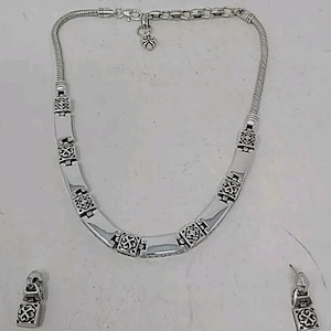 Puede incluir: Un conjunto de collar y pendientes plateados. El collar presenta una cadena de serpiente y una serie de eslabones rectangulares con un patr&oacute;n decorativo. Tambi&eacute;n se incluyen pendientes a juego, cada uno con un dise&ntilde;o similar. El conjunto de joyas se muestra sobre una superficie blanca.