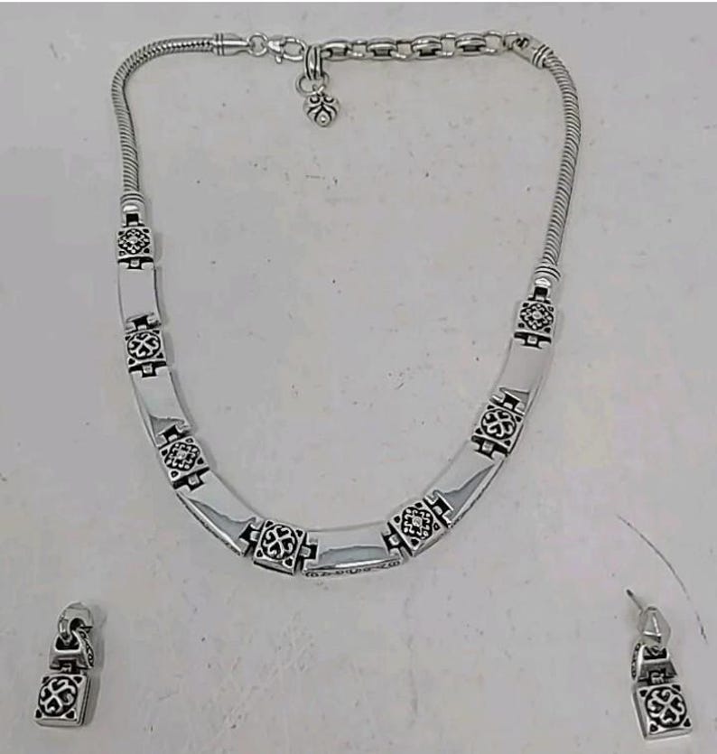 Puede incluir: Un conjunto de collar y pendientes plateados. El collar presenta una cadena de serpiente y una serie de eslabones rectangulares con un patr&oacute;n decorativo. Tambi&eacute;n se incluyen pendientes a juego, cada uno con un dise&ntilde;o similar. El conjunto de joyas se muestra sobre una superficie blanca.