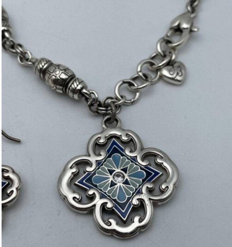 May include: A silver necklace and earrings set with a blue and white enamel pendant featuring a floral design. The pendant is surrounded by a silver filigree frame.