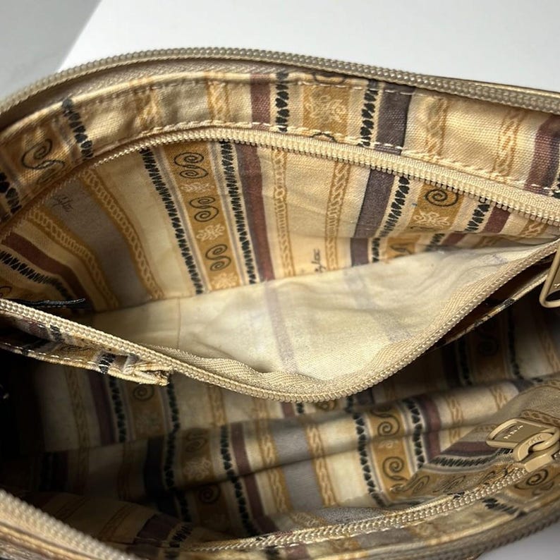 May include: Open view of a beige handbag interior. The inside features a patterned fabric with stripes, swirls, and geometric shapes in shades of brown and beige. A zipper runs along the top edge, and the bag appears to be empty.