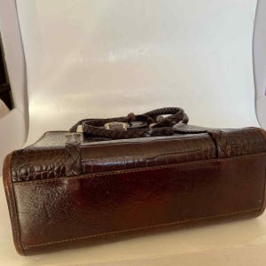 May include: A brown leather handbag with a crocodile-textured exterior. The bag features a braided handle and a silver-colored buckle closure. The corners of the bag are rounded, and the overall design is classic and elegant.