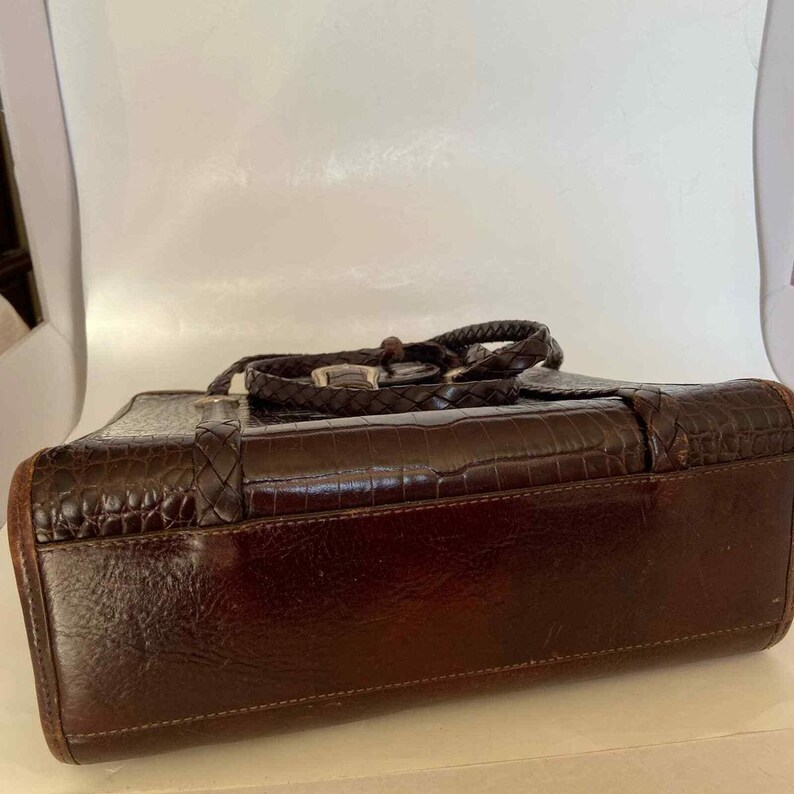 May include: A brown leather handbag with a crocodile-textured exterior. The bag features a braided handle and a silver-colored buckle closure. The corners of the bag are rounded, and the overall design is classic and elegant.