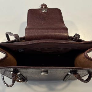 May include: Open view of a brown leather handbag with a textured crocodile pattern. The bag has a flap closure and braided handles. The interior is visible, revealing a spacious compartment.