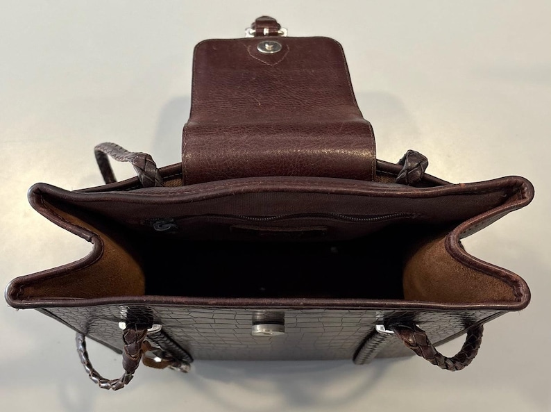 May include: Open view of a brown leather handbag with a textured crocodile pattern. The bag has a flap closure and braided handles. The interior is visible, revealing a spacious compartment.