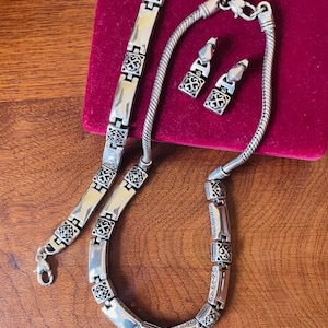 Puede incluir: Un conjunto de joyas plateadas que incluye un collar, una pulsera y pendientes. El collar y la pulsera tienen un dise&ntilde;o de eslabones con detalles cuadrados decorativos. Los pendientes son de estilo colgante a juego. El conjunto se exhibe sobre una superficie de terciopelo rojo.