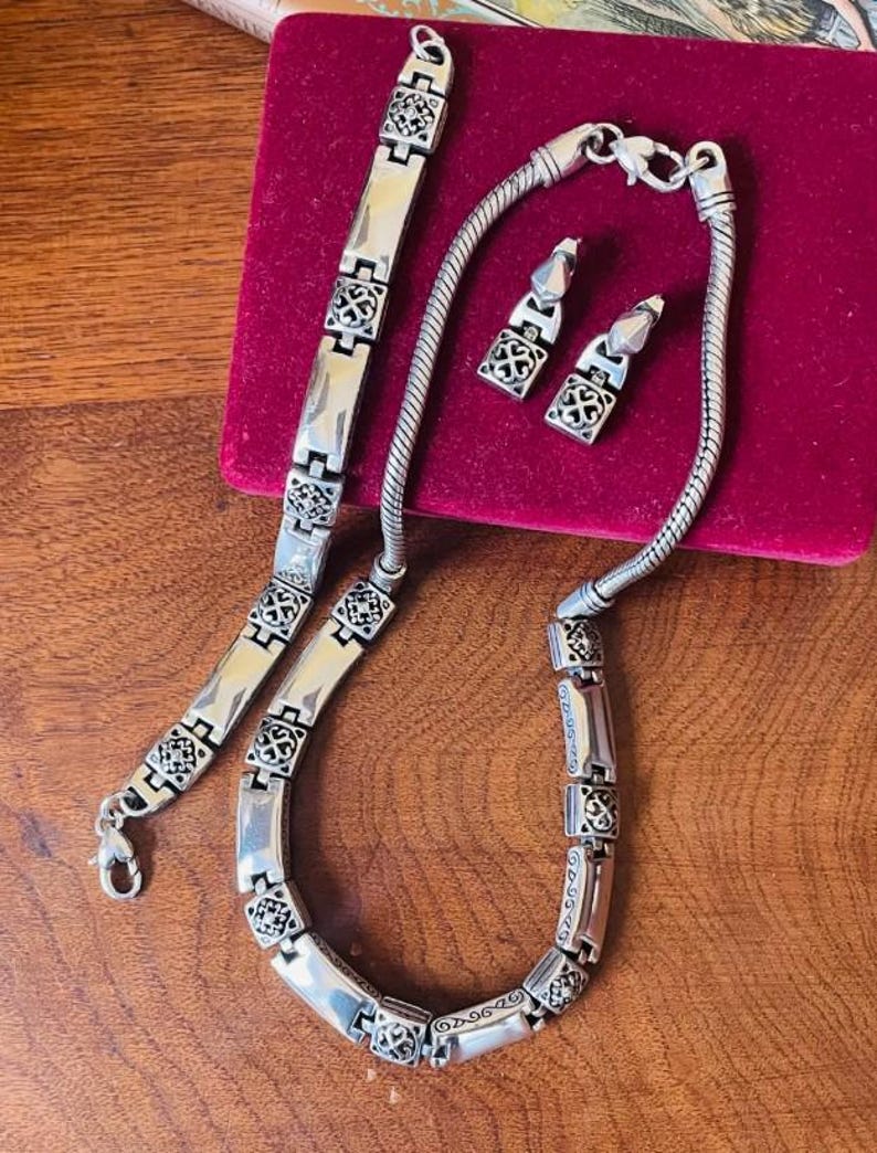 Puede incluir: Un conjunto de joyas plateadas que incluye un collar, una pulsera y pendientes. El collar y la pulsera tienen un dise&ntilde;o de eslabones con detalles cuadrados decorativos. Los pendientes son de estilo colgante a juego. El conjunto se exhibe sobre una superficie de terciopelo rojo.