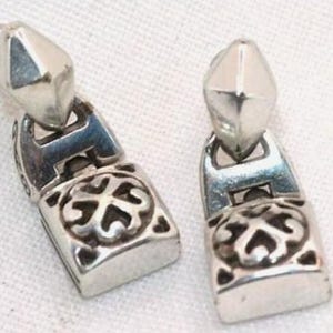 Puede incluir: Dos pendientes plateados. Cada pendiente presenta una parte superior geom&eacute;trica facetada y una base rectangular con un dise&ntilde;o ornamental y sim&eacute;trico. Los pendientes se muestran sobre una superficie blanca.