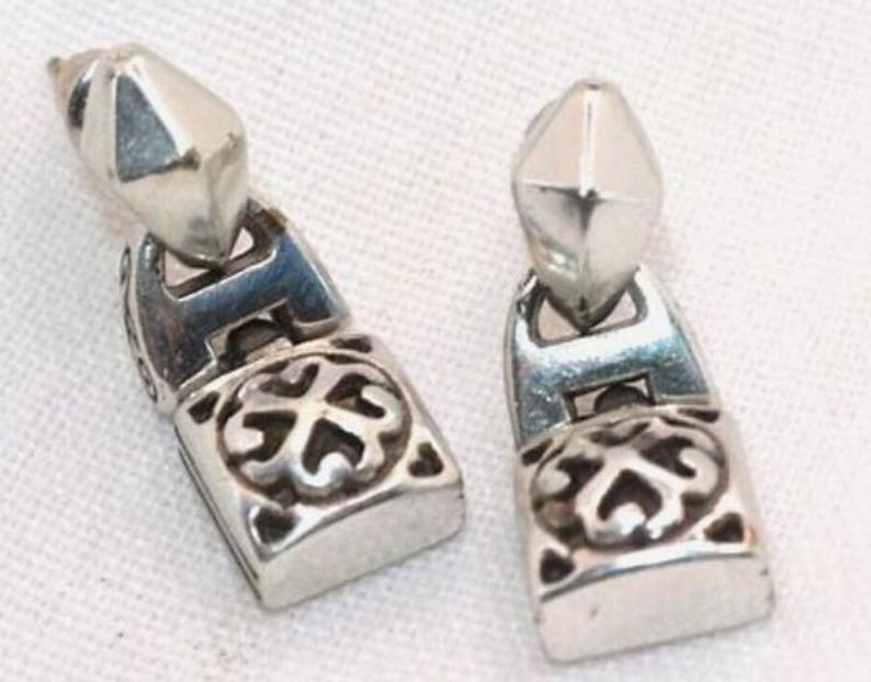 Puede incluir: Dos pendientes plateados. Cada pendiente presenta una parte superior geom&eacute;trica facetada y una base rectangular con un dise&ntilde;o ornamental y sim&eacute;trico. Los pendientes se muestran sobre una superficie blanca.