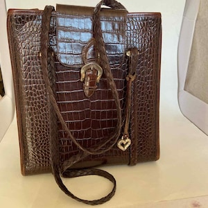 May include: A brown, faux-crocodile leather handbag with a braided shoulder strap. The bag features a flap closure with a silver-colored buckle and a small heart charm. The bag is rectangular and has a structured design.
