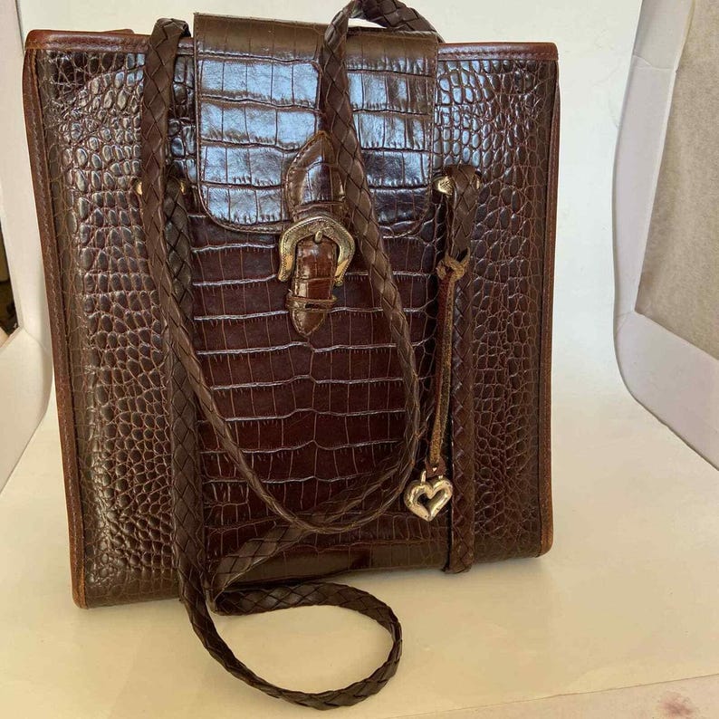 May include: A brown, faux-crocodile leather handbag with a braided shoulder strap. The bag features a flap closure with a silver-colored buckle and a small heart charm. The bag is rectangular and has a structured design.