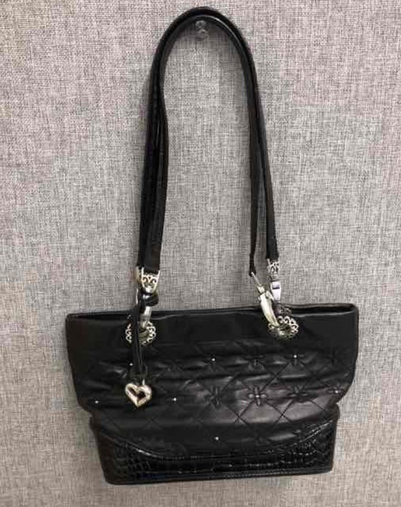 Pu&ograve; includere: Borsa tote in pelle trapuntata nera con dettagli in argento e un ciondolo a forma di cuore. La borsa ha una chiusura con cerniera superiore e una lunga tracolla.