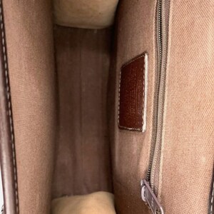 May include: Interior view of a brown leather handbag. The bag features a light brown fabric lining, a brown leather tag, and a silver zipper. The bag's interior is visible, showcasing its spacious design.