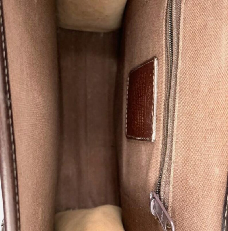 May include: Interior view of a brown leather handbag. The bag features a light brown fabric lining, a brown leather tag, and a silver zipper. The bag's interior is visible, showcasing its spacious design.