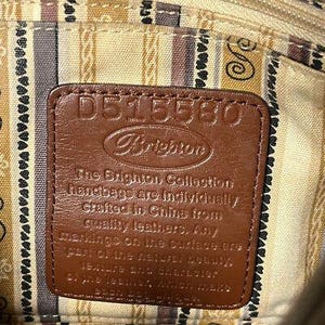 May include: Close-up of a brown leather tag with the Brighton logo and the number D515580. The tag is stitched onto a patterned fabric. The text on the tag reads "The Brighton Collection handbags are individually crafted in China from quality leathers."