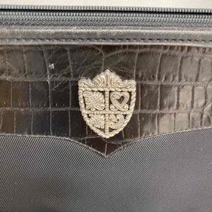 K&ouml;nnte beinhalten: Eine schwarze Lederhandtasche mit einem silbernen, verzierten, schildf&ouml;rmigen Verschluss.