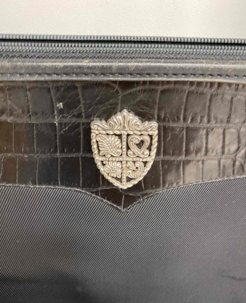 K&ouml;nnte beinhalten: Eine schwarze Lederhandtasche mit einem silbernen, verzierten, schildf&ouml;rmigen Verschluss.