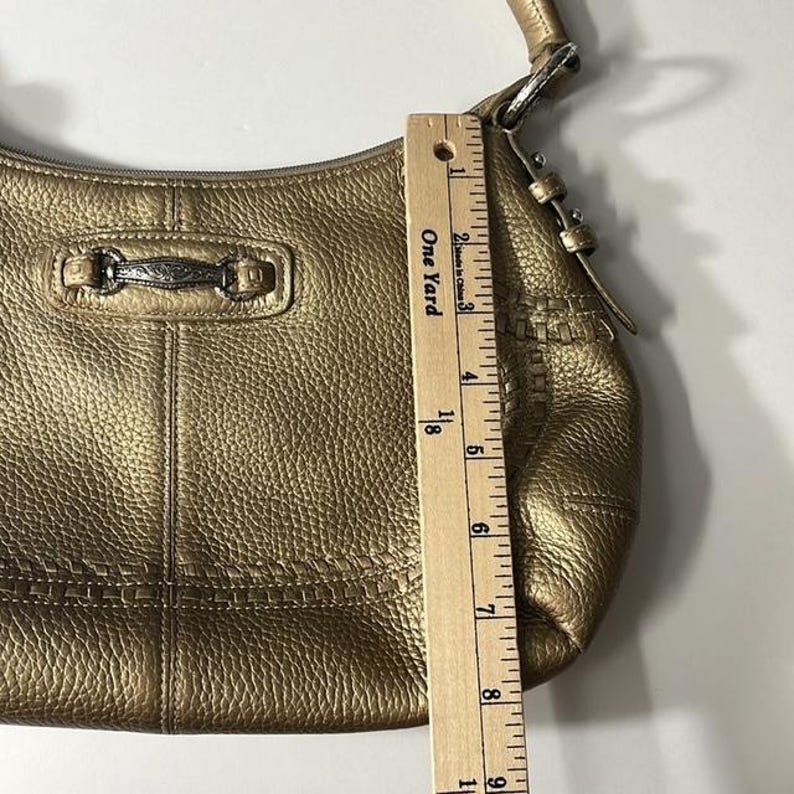 May include: A metallic gold-colored handbag with a textured finish. The bag has a curved shape with a top handle and a decorative metal clasp. The ruler shows the bag is approximately 9 inches long.