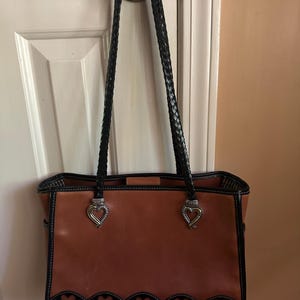 May include: A brown leather tote bag with black braided handles and silver heart-shaped accents. The bag features a black scalloped trim with heart cutouts along the bottom edge. White stitching outlines the bag's edges.