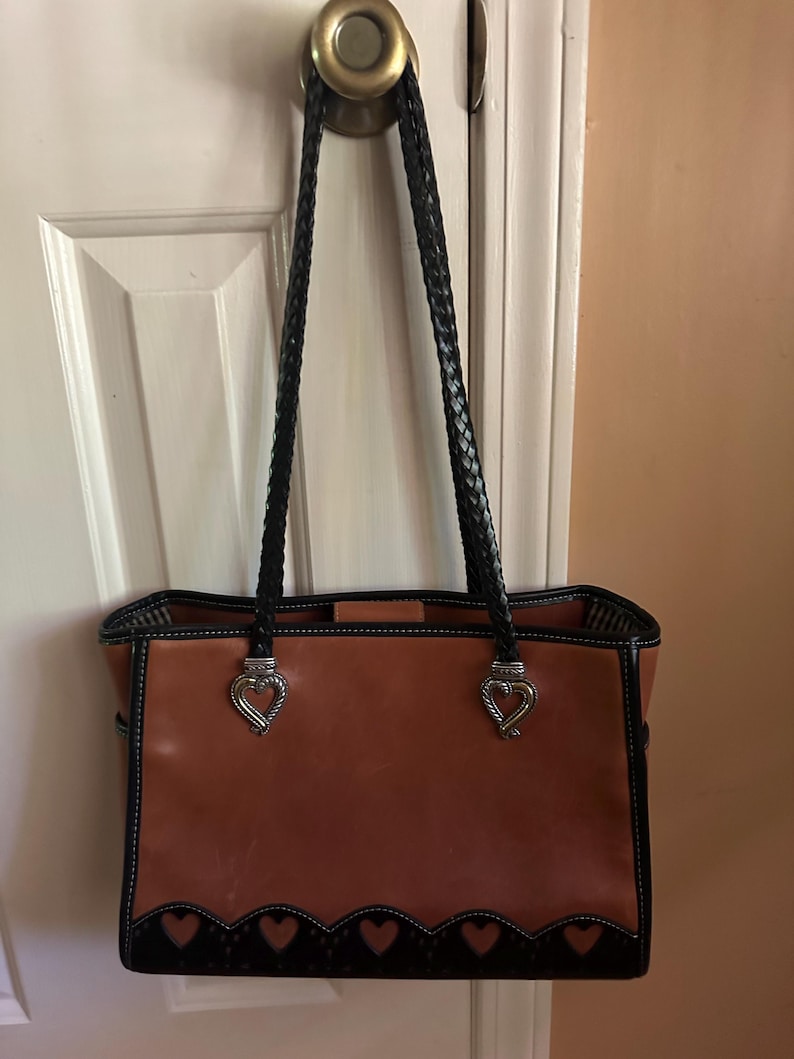 May include: A brown leather tote bag with black braided handles and silver heart-shaped accents. The bag features a black scalloped trim with heart cutouts along the bottom edge. White stitching outlines the bag's edges.