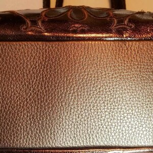 May include: A brown leather handbag with a textured surface. The top of the bag features a decorative, dark brown panel with an ornate design. The bag has a rounded shape and appears to be a shoulder bag.