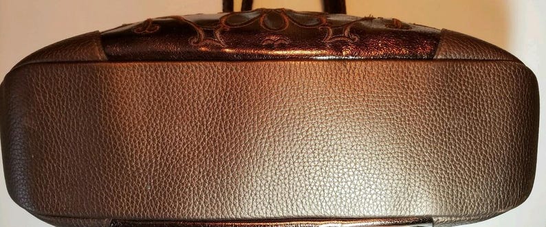 May include: A brown leather handbag with a textured surface. The top of the bag features a decorative, dark brown panel with an ornate design. The bag has a rounded shape and appears to be a shoulder bag.