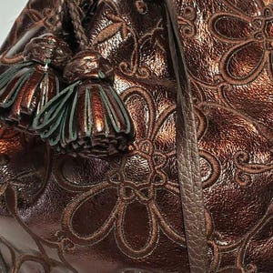 May include: Close-up of a brown leather handbag with a floral embossed pattern. The bag features a braided handle, decorative tassels, and a silver heart-shaped charm. The leather has a metallic sheen.