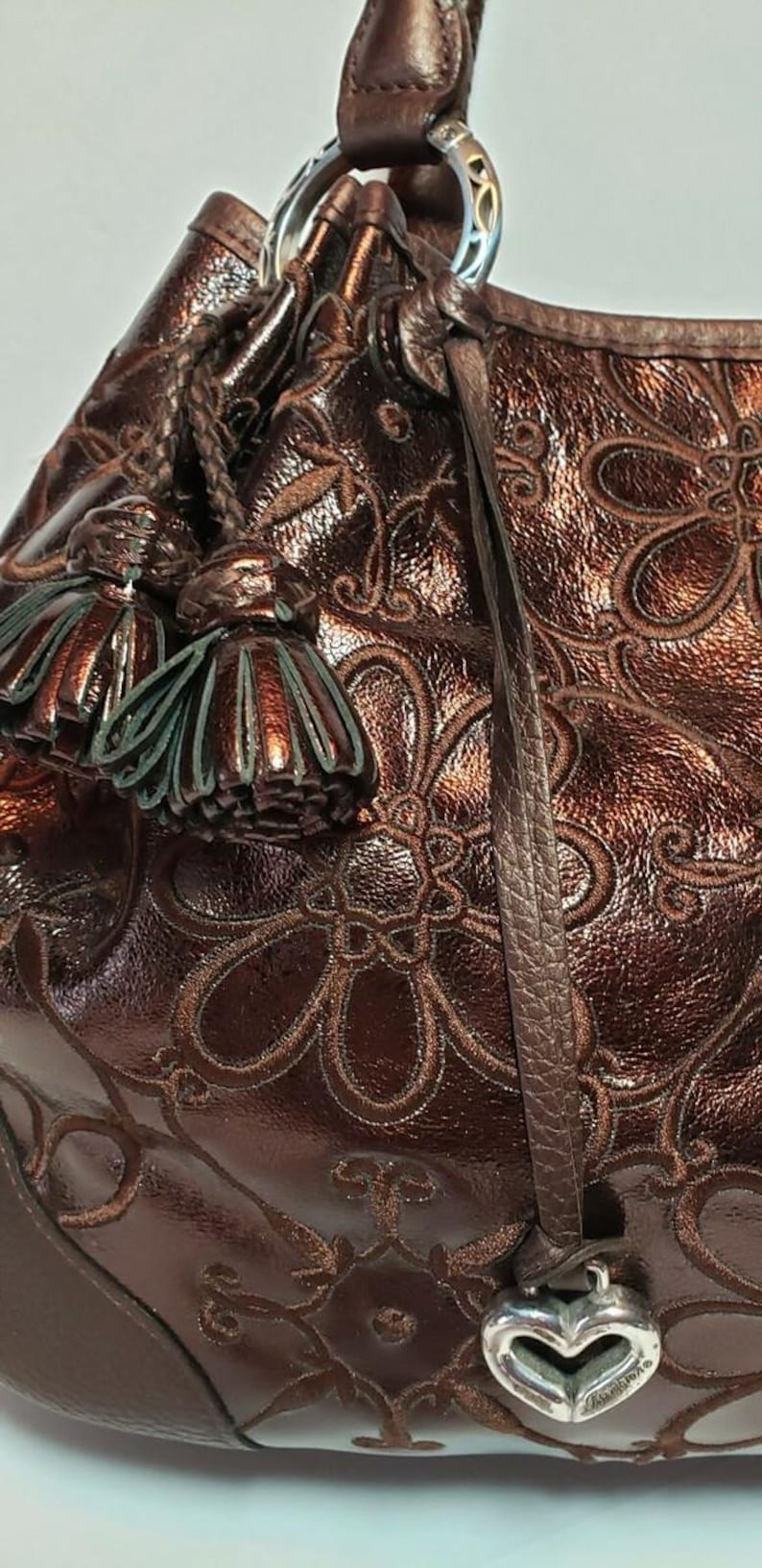 May include: Close-up of a brown leather handbag with a floral embossed pattern. The bag features a braided handle, decorative tassels, and a silver heart-shaped charm. The leather has a metallic sheen.