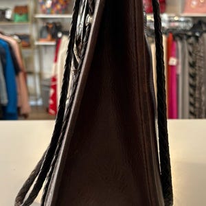 May include: A dark brown leather tote bag with braided handles. The bag has a rectangular shape and a textured surface. The handles are a darker shade of brown. The bag is displayed on a white surface.